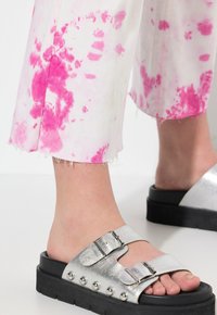 Silverfärgade metalliska slidesandaler med två justerbara remmar och nitar. Matchas med vita, rosa batikmönstrade cropped byxor med fransar.