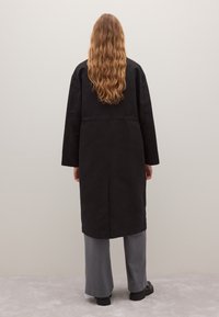 Langer schwarzer Mantel mit geradem Schnitt und langen Ärmeln, aus glattem Stoff gefertigt. Kombiniert mit hellgrauen, weit geschnittenen Hosen.