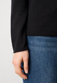 Schwarzer gestrickter Pulloverärmel mit geripptem Bund, kombiniert mit strukturierten blauen Jeans. Nahaufnahme einer Hand, die an der Naht ruht.
