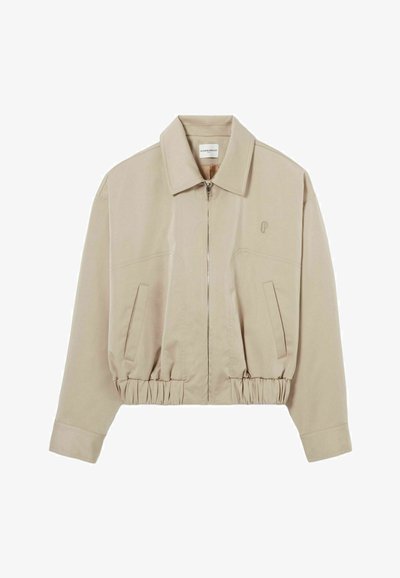 Giacca beige cropped con colletto a risvolto, chiusura con zip, orlo elasticizzato e due tasche frontali. Presenta un logo ricamato discreto sul petto.