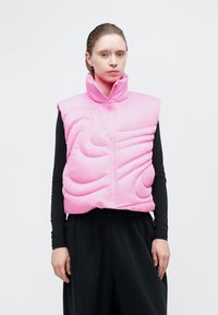Teksturirani ružičasti puffer prsluk s visokim ovratnikom i prednjim patentnim zatvaračem. Ima jedinstvene uzorke šavova i nosi se preko crnog long-sleeve majice.