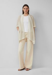 Cardigan léger en maille ouverte, de couleur crème, avec des manches courtes et un design drapé. Associé à un haut blanc et un pantalon clair.