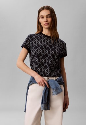Kvinne med brunt hår har på seg en svart mønstret topp med korte ermer, hvite bukser, og holder en blå denimjakke knytt rundt midjen, nøytral grå bakgrunn.