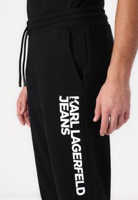 Melni sporta bikses ar aukliņu jostasvietā, ar baltu tekstu "KARL LAGERFELD JEANS" kreisajā pusē, mīksta auduma un sānu kabatām.