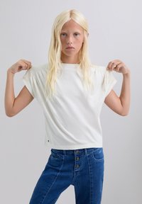 Top blanco de manga corta con detalle de hombro fruncido, combinado con jeans azul de tiro alto que cuentan con cierre de cinco botones y bolsillos laterales.