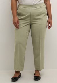 Pantalons ajustés vert clair avec une texture lisse, jambes fuselées, poches latérales et une taille plate, associés à des chaussures plates noires.
