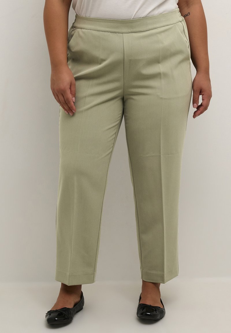 Pantalons ajustés vert clair avec une texture lisse, jambes fuselées, poches latérales et une taille plate, associés à des chaussures plates noires.