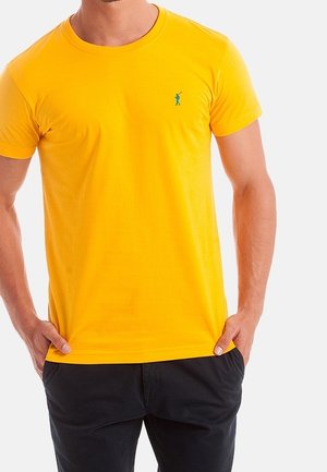Homme portant un t-shirt jaune vif à manches courtes avec un petit logo de golfeur vert et un pantalon noir, les mains dans les poches.