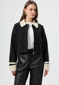 Zwarte cropped jas met een zachte, crèmekleurige kraag en boorden. Voorzien van een ritssluiting aan de voorkant en zakken, gecombineerd met zwarte nepleren broek.