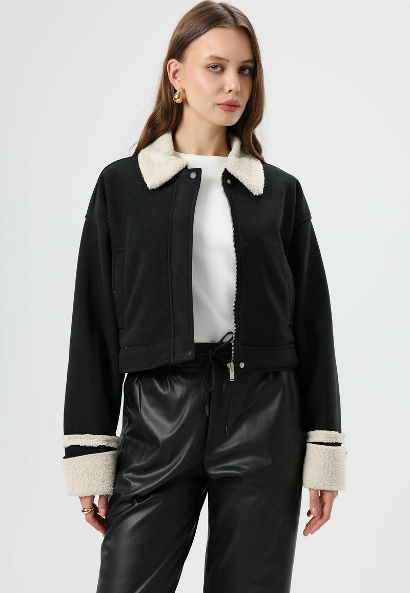 Zwarte cropped jas met een zachte, crèmekleurige kraag en boorden. Voorzien van een ritssluiting aan de voorkant en zakken, gecombineerd met zwarte nepleren broek.