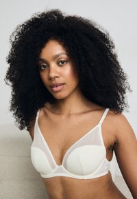 Crème-kleurige beha met zachte, gepolsterde cups, doorzichtige mesh zijpanelen en verstelbare banden. Eenvoudig, elegant ontwerp dat het comfort benadrukt.