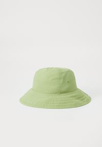 Un chapeau seau vert clair en tissu doux, avec une large visière et des détails cousus autour de la base. Il présente des trous de ventilation sur les côtés.