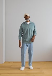 Lichtblauwe rits-hoodie, comfortabele pasvorm, lange mouwen. Gecombineerd met lichtblauwe jeans en witte sneakers, staande op een houten vloer.