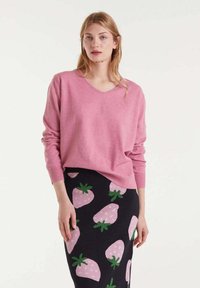 Suéter de punto de pico rosa con mangas largas, combinado con una falda oscura que presenta grandes patrones de fresas rosas y acentos verdes. Textura suave visible.
