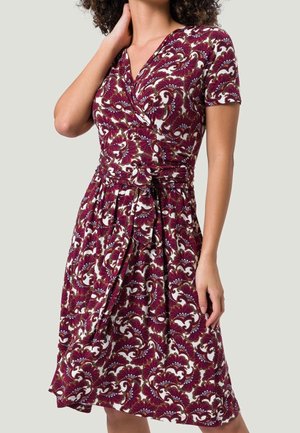 Frau trägt ein kurzes, knielanges Wickelkleid mit kurzen Ärmeln, einem floralen Muster in Burgunderrot, Weiß und Gold sowie einem gebundenen Taillengürtel.