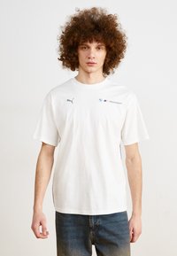 Camiseta de algodón blanca de manga corta, con cuello redondo, que presenta un logo gris de Puma y la marca BMW Motorsport en el lado izquierdo del pecho. Diseño sencillo.
