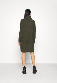 Strikkjorset i mørkegrøn med høj krave, lange ærmer og en tætsiddende silhuet, der når til midt på lårene, parret med sorte ankelstøvler.