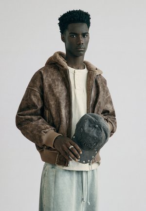 Jeune homme portant une veste en peau lainée marron et un pantalon clair tient une casquette de baseball noire usée avec des œillets argentés sur un fond gris uni.