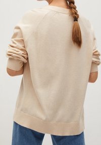 Beżowy sweter z prążkowanymi mankietami i dołem, luźny krój, z delikatną teksturą i rękawami raglanowymi, noszony na niebieskich dżinsach.