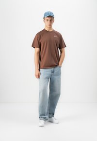 Young man wearing light blue cap, brown short-sleeve shirt, loose light blue jeans, and white sneakers, standing against white background.