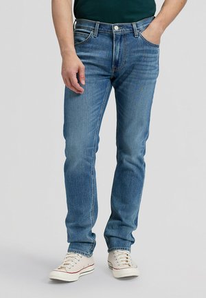 Mężczyzna w niebieskich jeansach typu slim-fit i białych tenisówkach stoi z jedną ręką w kieszeni na jednolitym tle.