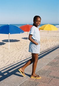Jeune fille en chemise bleu clair et short marche sur le trottoir près d’une plage de sable avec des parasols bleus, rouges et jaunes sous un ciel clair.