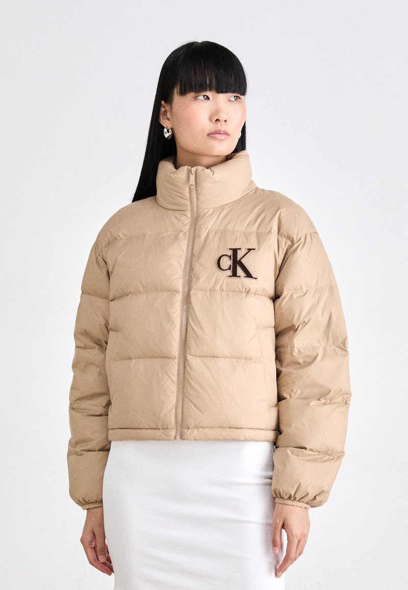 Doudoune beige avec un col montant, présentant des sections matelassées, une fermeture éclair et un logo "cK" marron sur la poitrine gauche.