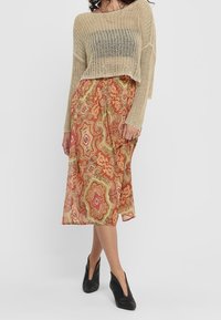 Une femme pose dans un pull en maille beige court et une jupe maxi colorée et à motifs, associée à des talons noirs, devant un fond uni.