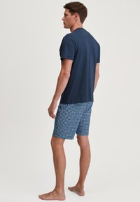 Jonge man die blootsvoets staat, gekleed in een marineblauw T-shirt en blauwe shorts met patroon, met zijn gezicht naar links tegen een effen achtergrond.