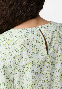 Blomstret bluse med en lyseblå baggrund og grønne accenter. Har en nøglehulåbning bagpå, sikret med en knap. Glat stof tekstur.