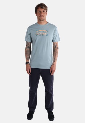 T-shirt en coton bleu clair avec un motif en orange et vert, associé à un pantalon bleu marine foncé et des chaussures noires. Détails avec des tatouages sur les bras.