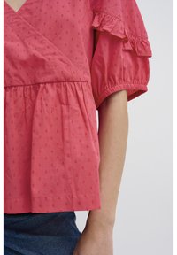 Blouse rose corail avec un décolleté en V, des manches courtes bouffantes et des accents à volants. Présente une taille froncée et un motif à pois texturé sur l'ensemble.