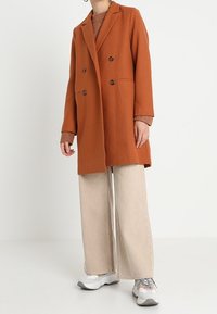 Abrigo de corte doble en mezcla de lana color naranja quemado, con botones marrones, solapas con muesca y bolsillos laterales, combinado con pantalones de pierna ancha en beige claro.