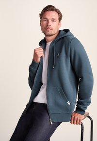 Teal zip-up hoodie in soft fabric with kangaroo pockets, ribbed cuffs, and a small logo on the chest, worn over a white t-shirt.