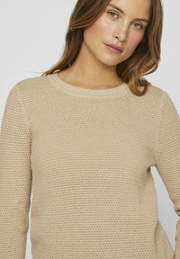 Bežani pleteni pulover s teksturiranim uzorkom, okruglim izrezom i dugim rukavima. Mekani, lagani materijal osigurava udoban kroj.