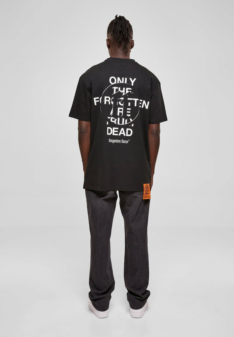 Juodas marškinėlis su ryškiai balta tekstu grafiniame dizaine nugaroje, derinamas su laisvai krentančiais tamsiais džinsais ir baltais sportbačiais.