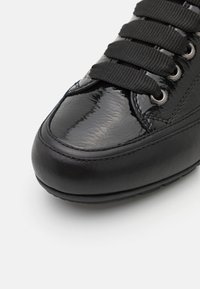 Schwarzer Sneaker mit glänzendem Lackleder an der Zehenkappe, strukturiertem Leder-Obermaterial und flachen schwarzen Schnürsenkeln. Rundliche Form mit Gummisohle.