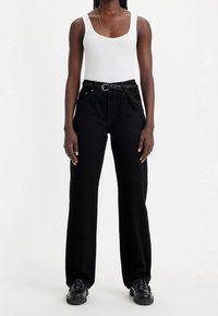 Vaqueros negros de tiro alto y pierna ancha con un cinturón trenzado, que incluyen bolsillos delanteros y un tejido liso y duradero. Combinados con una camiseta sin mangas blanca.