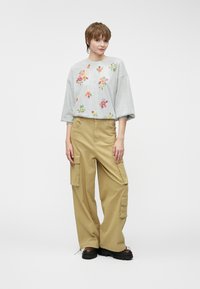 Sivý sveter s kvetinovou výšivkou, skombinovaný s khaki cargo nohavicami s bočnými vreckami. Model má na sebe robustné čierne topánky.