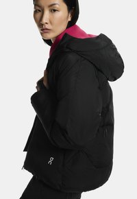 Fekete puffos dzseki kapucnival, rózsaszín bélésű, cipzáras zsebekkel és logós részlettel az oldalon.