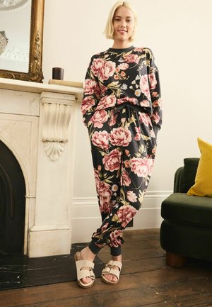 Floral loungewear set in dark navy, featuring large pink and cream blossoms. Loose fit, elastic waistband, and grey cuffs. Beige sandals with buckles.