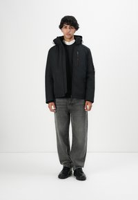 En ung mand iført en sort hættetrøje, en sort sweater, en hvid skjorte, falmede grå jeans og sorte sko, der står mod en ensfarvet hvid baggrund.