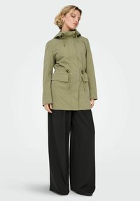 Parka kaki avec capuche et taille réglables par cordon, dotée de deux poches avant et d'une coupe droite, associée à un pantalon noir à jambes larges.