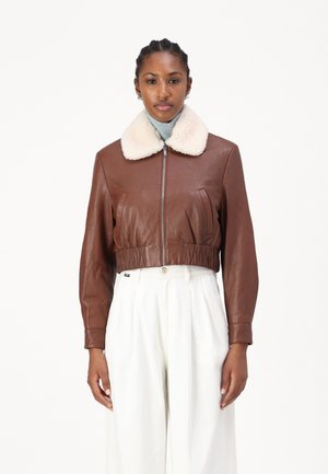 Kamelbraune Lederjacke mit einem weißen Schafwollkragen, Reißverschluss und elastischem Saum. Kombiniert mit hellen, weiten Hosen.