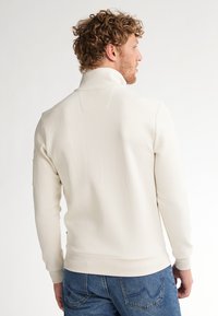 Pulover alb cu guler înalt, fabricat dintr-un material moale, având un design simplu, manșete cu nervuri și un detaliu de yoke la spate. Asortat cu blugi albaștri.