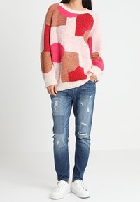 Přoversize růžový svetr s mnohobarevnými geometrickými vzory, pletená textura; spárovaný s roztrhanými modrými džíny a bílými teniskami.