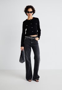 Black cropped cardigan with button closure, paired with high-waisted, flared dark denim jeans. Accessories include a circular belt and bag.