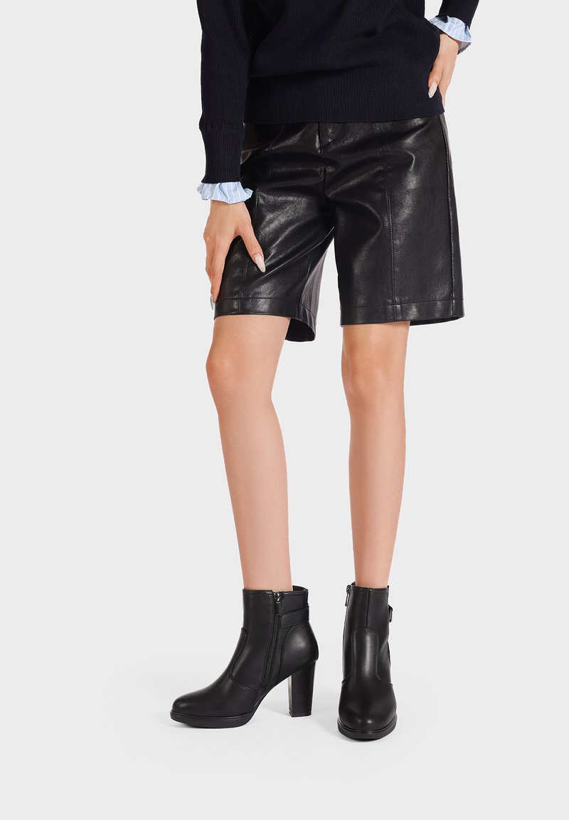 Stivali neri in pelle con tacco alto e cerniera laterale, abbinati a pantaloni shorts neri in pelle e un maglione blu navy con polsini arricciati azzurri.