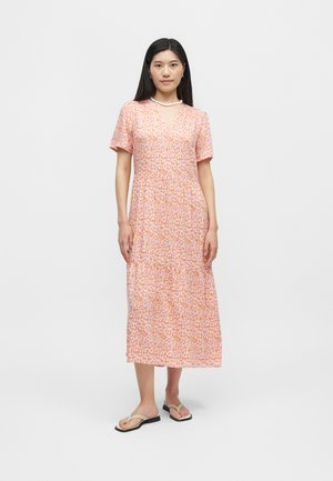 Žena stojaca v midi šatách s ružovo-oranžovým vzorom, s bielym náhrdelníkom a béžovými plochými sandálmi, na jednoliatom sivom pozadí.