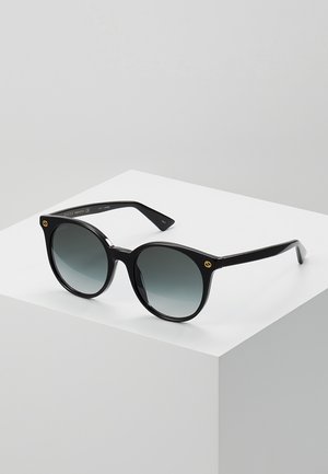 Lunettes de soleil noires avec une finition brillante, verres ronds passant d'un gris foncé à un gris clair, avec des accents dorés sur les branches.